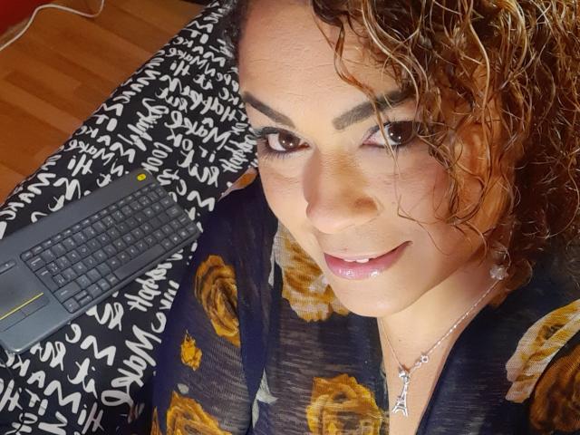 SolmaryHot with curly brown hair, brown eyes, and a slight smile, wearing a floral black shirt, has a laptop on her right, and a silver necklace, with a small drop.