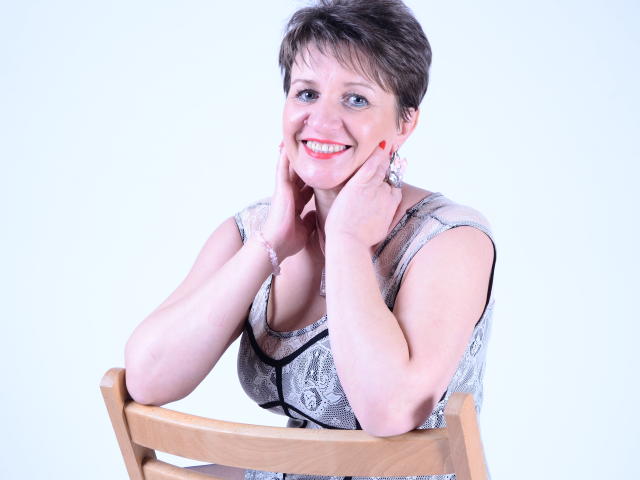 DoraBlueEyess with short brown hair, smiling, hands on cheeks, wearing a patterned gray top, sitting on a wooden chair, red nail polish, white background.