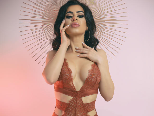LauraineX con senos de tamaño medio, vestida con lencería roja de encaje, con una mano en la mejilla, cabello oscuro y ondulado, aros grandes, tocado con forma de sol, fondo rosa y blanco. Pose suave y seductora..
