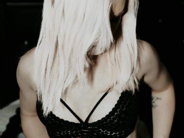 DreamArinna with platinum blonde hair, black lace bralette, and visible tattoos on her right arm, showing her tits and upper body. Her head is tilted down.