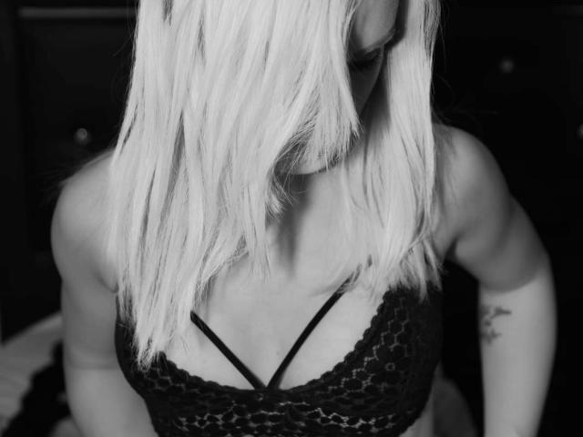 DreamArinna with platinum blonde hair covering her face, wearing a black lace bra with strappy details, showing her tits and a tattoo on her right arm. Black-and-white photo.