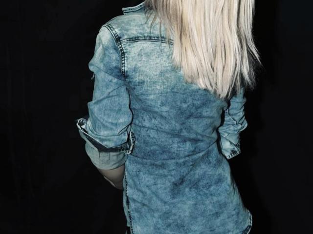 DreamArinna con i capelli biondi, indossa una camicia di jeans blu, leggermente sbiadita, è in piedi con la schiena rivolta verso la fotocamera, sfondo nero, mani nelle tasche, polsini della camicia arrotolati..