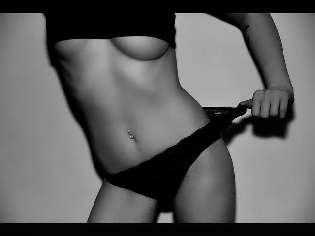 Schwarz-weißes Foto von DreamArinna mit flachem Bauch, die schwarze Spitzenunterwäsche hochhebt und dabei einen Bauchnabelpiercing sowie kleine, pralle Brüste zeigt. Ihr rechter Arm ist teilweise sichtbar..