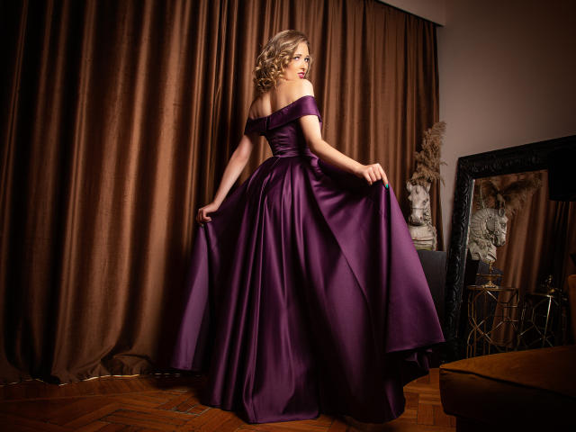 JessicaGibson in a purple off-shoulder ball gown, holding out the skirt, showing her ass, with curly blonde hair, standing in a dimly lit room.