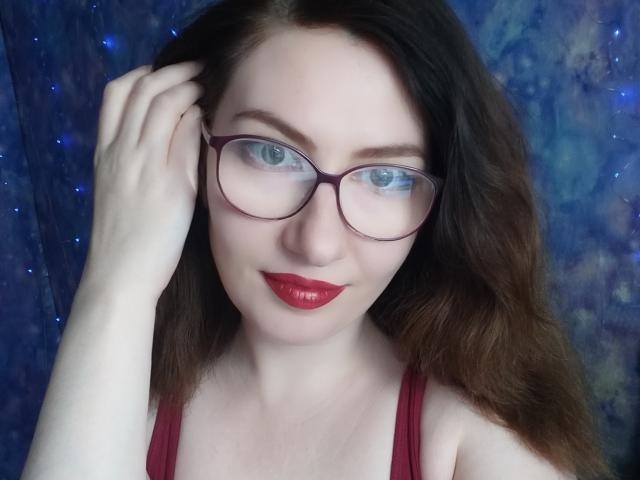 PaulinaSunny with red lipstick, glasses, and wavy brown hair, touching her head with pale skin and blue eyes, wearing a red tank top, showing her tits slightly.