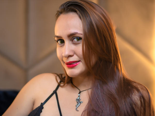 Katie Rilse, com cabelos castanhos longos, olhos verdes e batom vermelho, vestindo uma blusa preta de renda e um colar de cruz de prata, sorrindo levemente. Seus seios estão parcialmente visíveis..