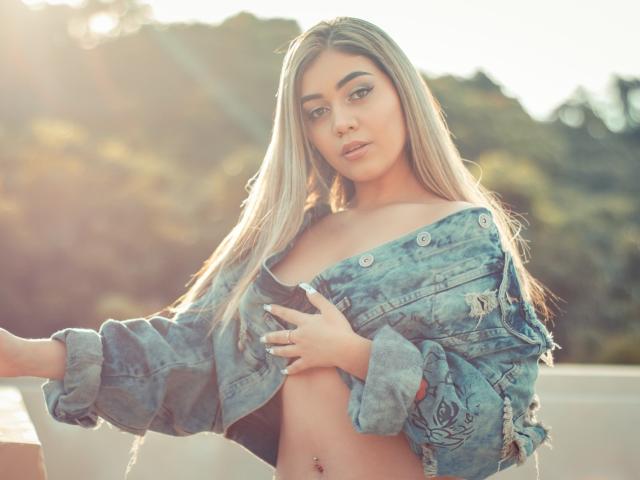 MiaMorton, avec de longs cheveux blonds, portant une veste en jean bleu ouverte, laissant entrevoir ses petites seins et un piercing au nombril, se tient au soleil et regarde l'appareil photo..