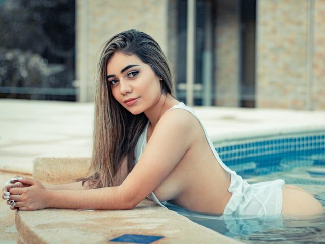 Mia Morton, con lunghi capelli castani, sdraiata in una piscina, indossa un costume intero bianco che rivela il seno e i glutei, e guarda la fotocamera con un leggero sorriso..
