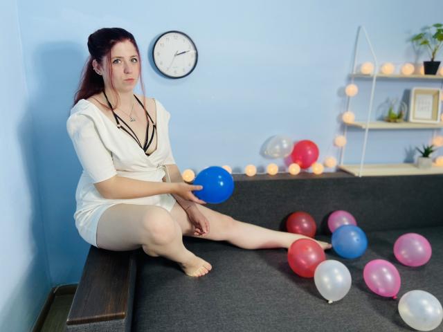 Jessika Daniels, avec sa peau pâle et ses cheveux roux, assise sur un canapé gris, tenant un ballon bleu, vêtue d'une robe blanche avec des détails noirs, entourée de ballons colorés..
