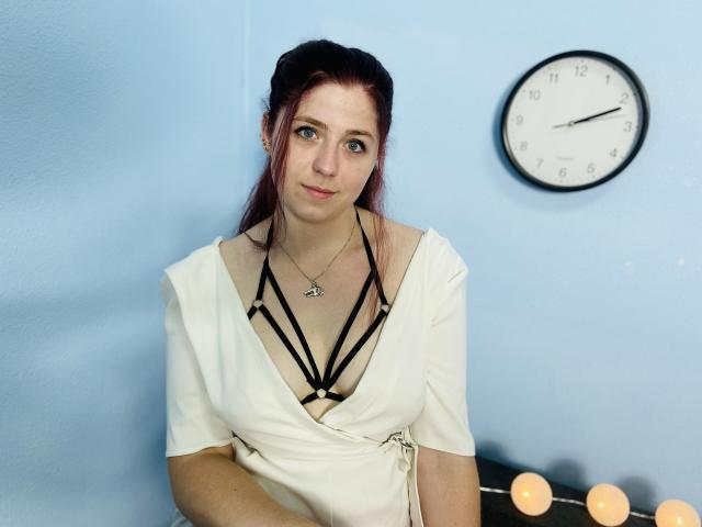 JessikaDaniels a les cheveux roux, les yeux bleus et un joli sourire. Elle porte un haut blanc avec de la lingerie noire à bretelles fines, laissant entrevoir ses seins. Une horloge est fixée sur un mur bleu clair..