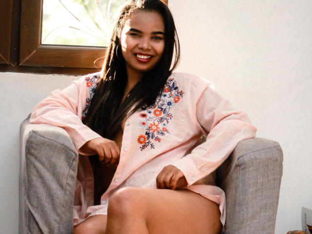 Charnelles with long dark hair, brown skin, and a smile, wearing a pink floral button-up shirt, sitting in a gray chair, sunlight through a window. Her pussy is visible.