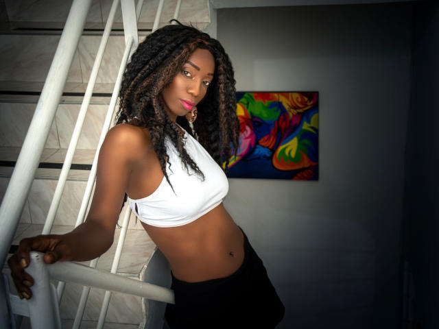 QueenLysse with dark brown skin, curly black hair, pink lipstick, white crop top, black pants, standing by a white staircase, colorful abstract painting in the background.