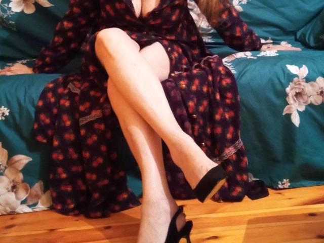 CarlyTreat's legs crossed, wearing a dark floral dress with a low neckline, black high heels, sitting on a teal floral couch, wooden floor, pale skin.