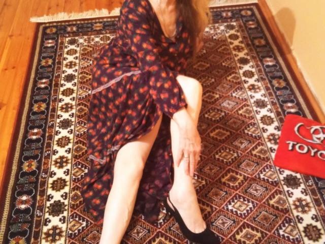 CarlyTreat, vestita con un abito floreale scuro, è seduta su un tappeto a motivi e sta tirando su l'abito per mostrare le sue gambe chiare e nude, con dei tacchi alti neri..