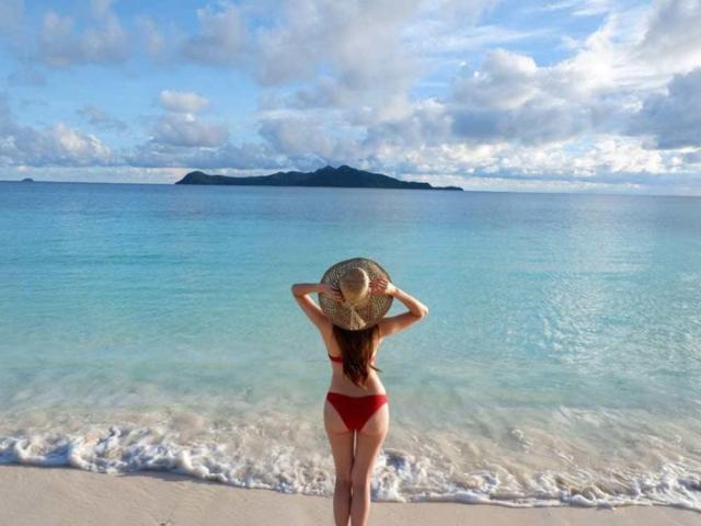 BeautyQueenTs staat op een strand, met haar gezicht naar de oceaan gericht, gekleed in een rood bikinie en een strohoed, met haar handen op de hoed, waarbij haar achterkant zichtbaar is, met helderblauw water en een bewolkte lucht..
