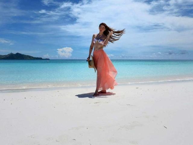 BeautyQueenTs with long brown hair, wearing a floral bra and pink sheer skirt, standing on a white beach, holding a straw bag, blue ocean, and clear sky.