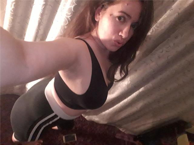 GoldySquirt, avec une poitrine de taille moyenne, un soutien-gorge de sport noir et un short de sport noir taille haute, tenant un téléphone pour un selfie, debout dans une pièce avec des rideaux beige..