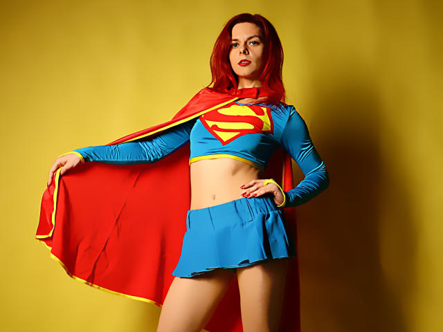 MelanyTessk with red hair, blue crop top, yellow trim, and red Superman emblem, red skirt, and red cape, standing against yellow background, hands on hips, confident.
