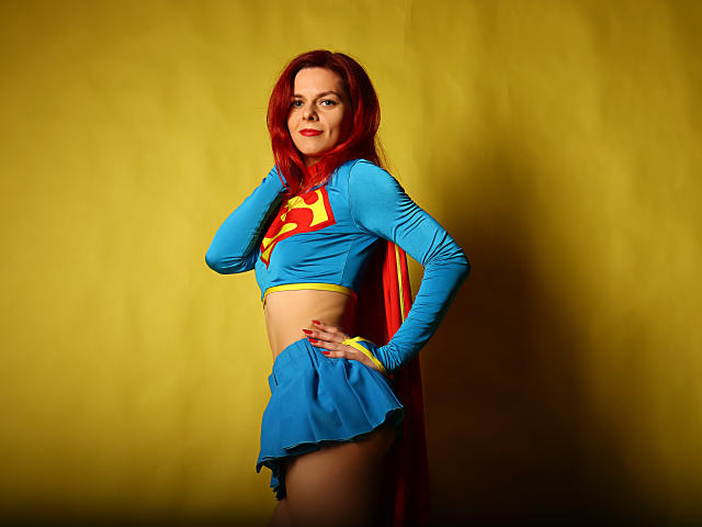 MelanyTessk vestida como Supergirl, con una blusa azul con la letra "S" amarilla y una falda mini roja y azul, una capa roja, cabello rojo, de pie con una mano en la cadera, sonriendo, fondo amarillo..