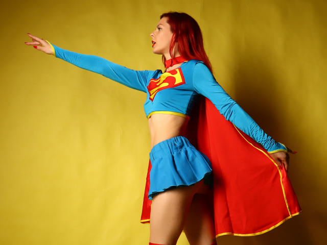 MelanyTessk in a Supergirl costume with blue top, red and blue skirt, and red cape, pointing to the left. Her tits are small, her ass is cute.