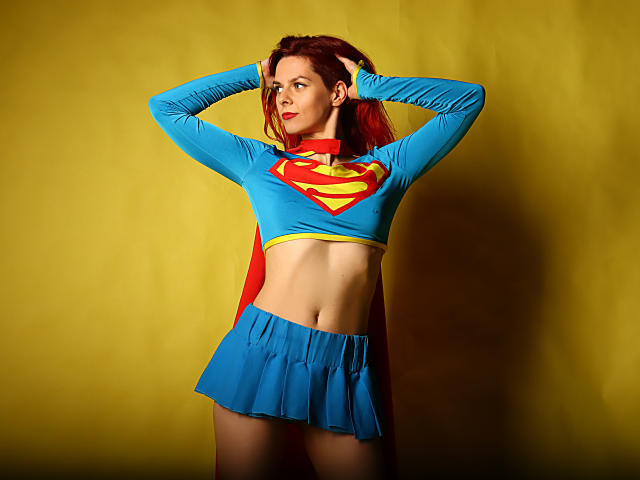 MelanyTessk in a Supergirl costume, blue top with red and yellow Super emblem, short blue skirt, red cape, hands in red hair, yellow background, slim, toned, red lipstick.