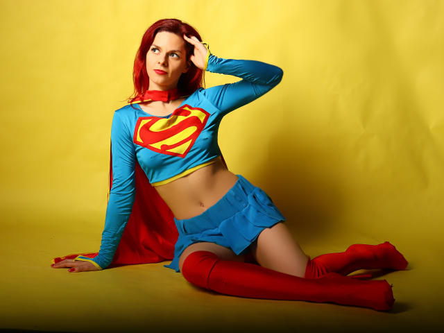 MelanyTessk with red hair, blue Supergirl crop top, red skirt, red boots, and red cape, sitting on yellow floor, hand on head, looking up, showing her ass.