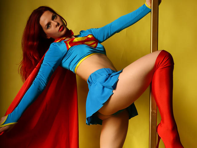 MelanyTessk in a Supergirl costume, with red boots and cape, bending over a pole, showing her pussy, tits, and toned midriff, with red hair and yellow background.