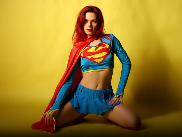 MelanyTessk, con cabello rojo, top corto azul con el logo de Superman, capa roja, falda mini azul, arrodillada, manos en los muslos, fondo amarillo, mirando a la cámara, plano medio..