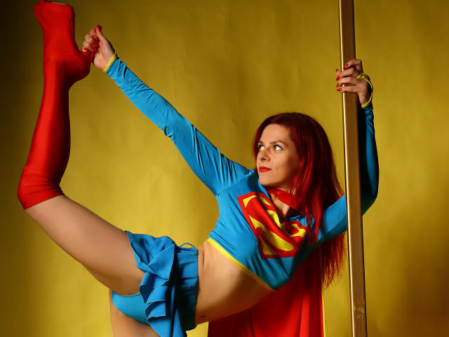 MelanyTessk, dressed as Superwoman, has red socks, blue crop top, and mini skirt, holding a pole, showing her ass, and raising one leg, with red hair.