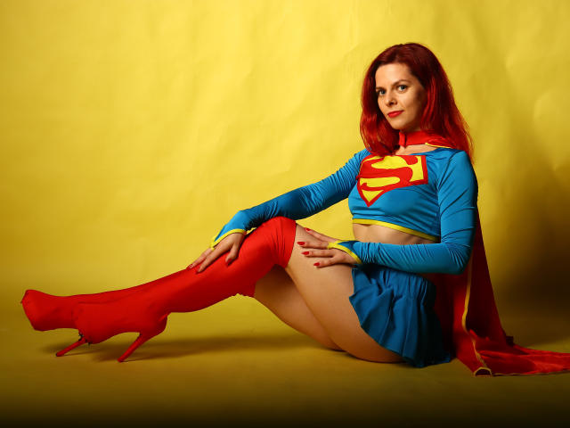 MelanyTessk is sitting with her legs out, wearing a Supergirl outfit with red boots, blue top, and yellow trim, showing her ass, with red hair and a cape.