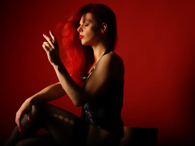 MelanyTessk with bright red hair, wearing black lace top and fishnet stockings, sits against a red background, flashing a peace sign, eyes closed, red lipstick, sexy.