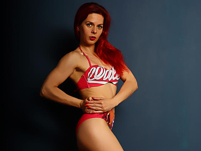 MelanyTessk, avec ses cheveux roux, portant un bikini rouge Coca-Cola, les mains sur son ventre plat, debout devant un mur bleu foncé, regardant l'appareil photo avec une expression sérieuse..
