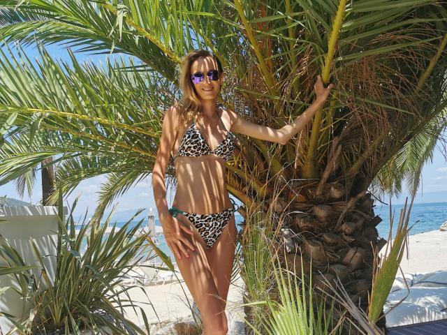 PlayfulErica stands in front of a palm tree, wearing a leopard print bikini, purple sunglasses, and smiling. She has medium tits and a slim, tan body. Beach background.