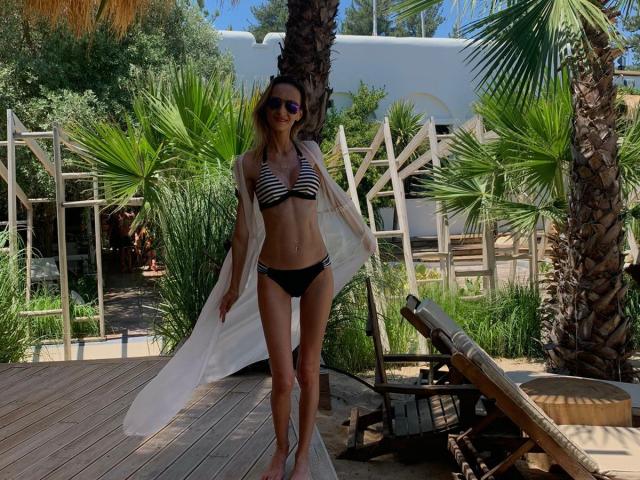 PlayfulErica with striped bikini top, black panties, and white sheer robe, standing on wooden deck, palm trees, sunglasses, long legs, slim, and small tits, next to lounge chair.