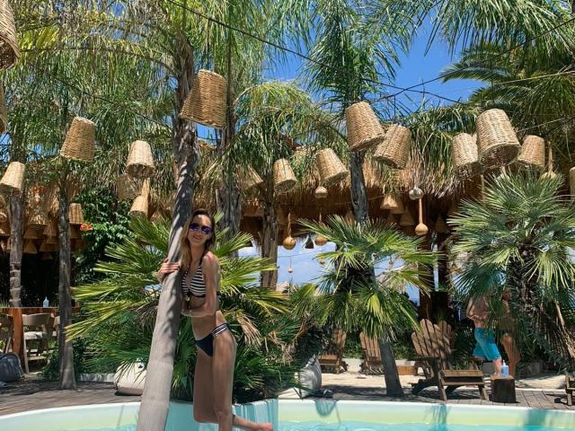PlayfulErica with medium tits in a black-and-white striped bikini, standing in a pool, holding a palm tree, wearing blue sunglasses, under hanging wicker lamps. Tropical setting.