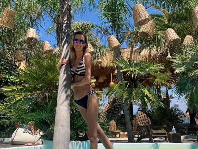 PlayfulErica with long brown hair, wearing a striped black-and-white bikini top and black bottoms, standing by a palm tree, smiling, wearing purple sunglasses, by a blue pool.