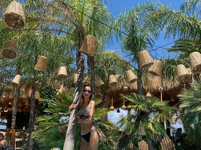 PlayfulErica in einem schwarz-weiß gestreiften Bikini, die einen Pfosten hält und unter hängenden Korblampen steht, trägt lila Sonnenbrillen, im Hintergrund sind Palmen und ein blauer Himmel zu sehen..