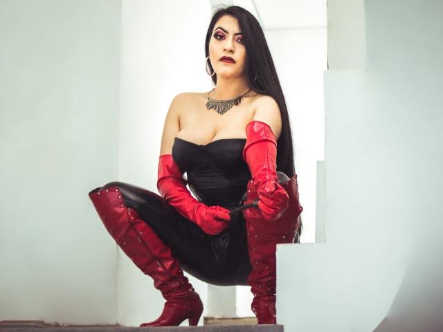 KamileNilsen está en posición de sentadilla, vistiendo un top negro sin tirantes, guantes de cuero rojo y botas a juego, con cabello negro largo, maquillaje rojo y un collar negro, mostrando sus pechos y glúteos..