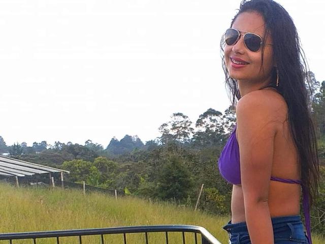 LeilaEvanns hat langes, dunkles, feuchtes Haar, trägt einen violetten Bikini-Oberteil, blaue Jeans und eine Sonnenbrille, lächelt mit rotem Lippenstift und steht neben einem Metallgeländer, im Freien..