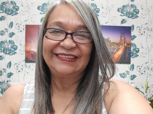 RosarioTriana tem cabelos acinzentados e castanhos, usa óculos, batom vermelho e tem um rosto sorridente com rugas visíveis, vestindo uma camiseta branca, mostrando suas nádegas na foto..