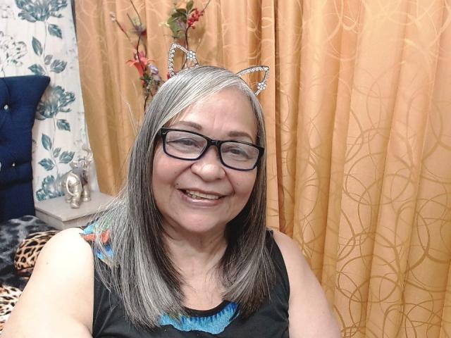Rosario Triana, com cabelos grisalhos, orelhas de gato, óculos pretos, sorrindo, vestindo uma blusa preta com renda azul, mostrando os dentes. Ela é uma mulher de meia-idade com uma aparência adorável..