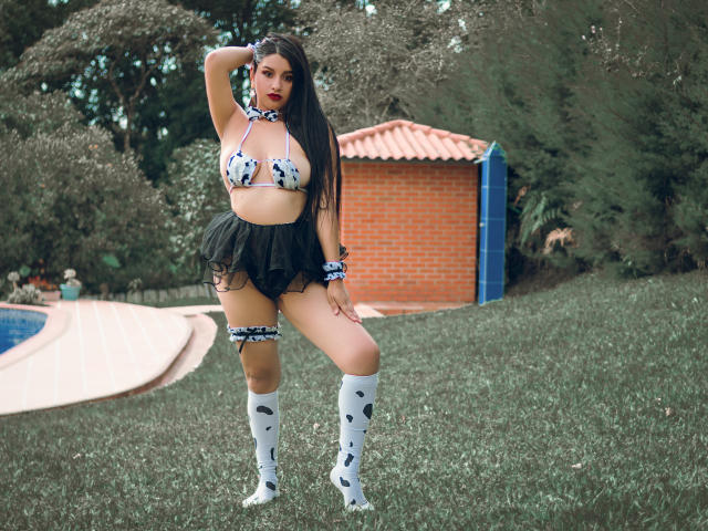SharolXD has big tits in a cow print bikini top, black mini skirt, white knee socks with black spots, and blue cow print accessories, standing on grass.