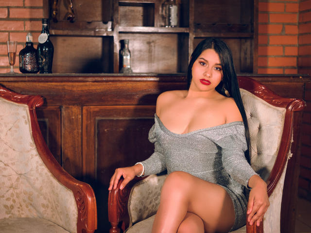 SharolXD with big tits, wearing a sparkly grey off-shoulder dress, sitting on a wooden chair, legs crossed, dark long hair, red lipstick, brick wall background, bottles on shelf.