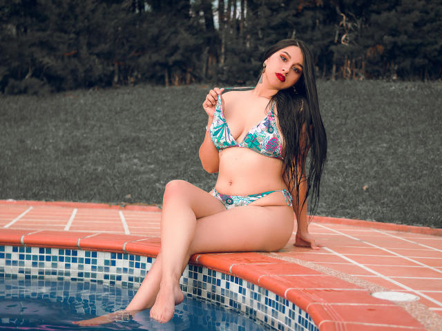 SharolXD with long black hair, wearing a floral bikini, sitting by the pool, holding a strap, showing her tits and ass, red lipstick, and a sexy, confident look.