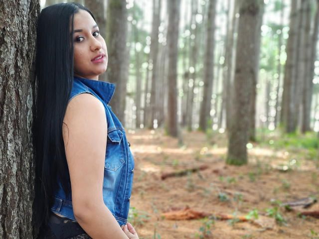 SharolXD with long black hair, denim sleeveless vest, standing against tree in forest, looking over shoulder, showing cute face and subtle smile, showing small, round ass.