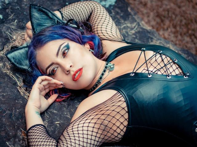 HannaTyllor mit blauen und lila Haaren, Katzenohren, schwarzen Netzstrümpfen, rotem Lippenstift, einem schwarzen Lederoberteil und einem Schnürdetail am Brustbereich, liegt auf dunklem Fels und blickt verführerisch in die Kamera..