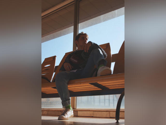 LuxMiamoto, dans une posture détendue et affalée, est assise sur un banc en bois, vêtue d'une veste verte, d'un jean bleu et de baskets blanches, baignée de lumière du soleil par derrière, les yeux à moitié ouverts, avec une expression d'ennui..