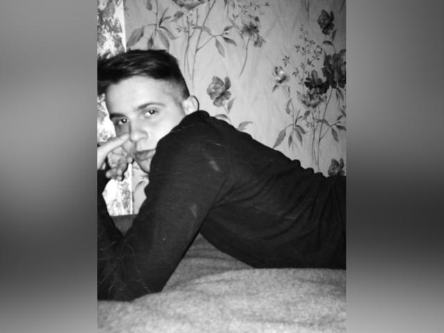 Fotografía en blanco y negro de LuxMiamoto acostada en una cama, vistiendo una camisa de manga larga oscura, con cabello corto y oscuro, mirando por encima del hombro, con un dedo cerca de la boca..