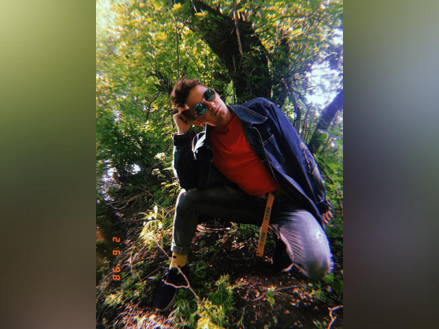LuxMiamoto accroupi dans une forêt, portant des lunettes de soleil, une chemise rouge, une veste noire, un jean gris, des chaussettes jaunes, des chaussures noires, tenant un mètre ruban jaune, avec des arbres verts luxuriants en arrière-plan..