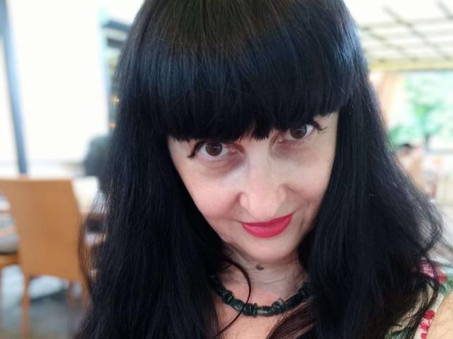 Melany Banks hat schwarzes, glattes, schulterlanges Haar mit Pony, braune Augen, roten Lippenstift und eine schwarze Perlenkette. Ihr Gesicht zeigt ein leichtes Lächeln, und sie hat einen kleinen Muttermal am Kinn..